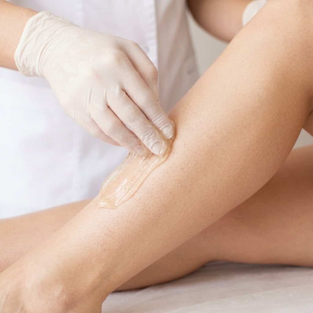 An esthetician's gloved hand precisely extracts hair using translucent sugar paste on a client's leg.